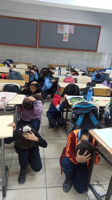 First image shows a classroom with young girls in colorful uniforms and headscarves sitting around a table covered with a white cloth some wearing animal-themed costumes like cow patterns others with backpacks on chairs bulletin boards with drawings and educational posters on walls red-trimmed displays and hanging clothes. Second image depicts students mostly boys and girls in blue uniforms kneeling or sitting on the floor in a classroom with desks pushed aside heads covered with arms or cloths simulating hiding during an earthquake gray board with posters water bottles and backpacks scattered around. Third image captures multiple students in similar uniforms crouching under or beside desks in a spacious classroom tiled floor gray board with decorations backpacks and water bottles on desks some children covering heads with arms or cloths during the drill exercise.