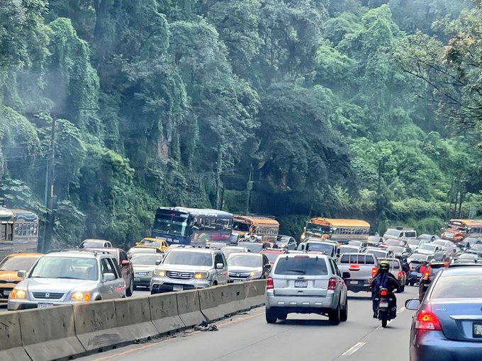 First image displays a line of yellow and white buses along with cars and a motorcycle on a multi-lane road bordered by dense green trees and hills under overcast skies with visible exhaust smoke. Second image shows heavy traffic including buses vans cars and a motorcycle with a rider wearing a yellow helmet navigating through the congestion on a road with green foliage and a light pole in the background. Third image captures similar congestion with buses cars and a motorcycle rider in a black jacket moving amid vehicles on the roadside with lush green surroundings and a concrete barrier.