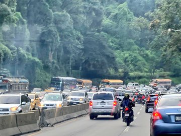 First image displays a line of yellow and white buses along with cars and a motorcycle on a multi-lane road bordered by dense green trees and hills under overcast skies with visible exhaust smoke. Second image shows heavy traffic including buses vans cars and a motorcycle with a rider wearing a yellow helmet navigating through the congestion on a road with green foliage and a light pole in the background. Third image captures similar congestion with buses cars and a motorcycle rider in a black jacket moving amid vehicles on the roadside with lush green surroundings and a concrete barrier.