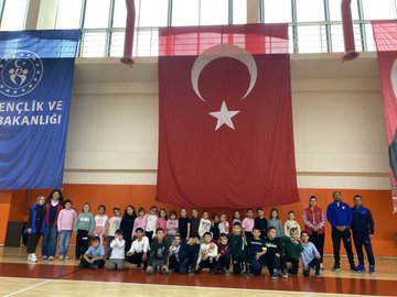 First image shows group of children holding certificates standing with adult male in suit in gymnasium with large Turkish flags and portrait of Mustafa Kemal Ataturk on walls. Second image depicts larger group of children and adults posing together in sports hall with banners for Ministry of Youth and Sports and Turkish flag. Third image captures children in circle playing game with ropes around central marker on basketball court wearing colorful clothing and some in headscarves. Fourth image features adult male and female with group of young girls holding hands near volleyball net in gym with bleachers in background.
