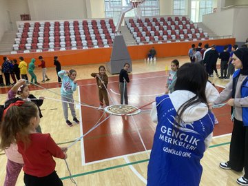 First image shows group of children holding certificates standing with adult male in suit in gymnasium with large Turkish flags and portrait of Mustafa Kemal Ataturk on walls. Second image depicts larger group of children and adults posing together in sports hall with banners for Ministry of Youth and Sports and Turkish flag. Third image captures children in circle playing game with ropes around central marker on basketball court wearing colorful clothing and some in headscarves. Fourth image features adult male and female with group of young girls holding hands near volleyball net in gym with bleachers in background.