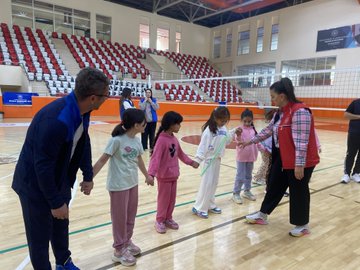 First image shows group of children holding certificates standing with adult male in suit in gymnasium with large Turkish flags and portrait of Mustafa Kemal Ataturk on walls. Second image depicts larger group of children and adults posing together in sports hall with banners for Ministry of Youth and Sports and Turkish flag. Third image captures children in circle playing game with ropes around central marker on basketball court wearing colorful clothing and some in headscarves. Fourth image features adult male and female with group of young girls holding hands near volleyball net in gym with bleachers in background.