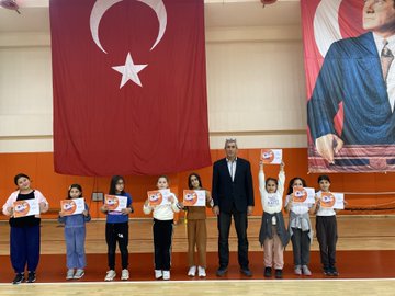 First image shows group of children holding certificates standing with adult male in suit in gymnasium with large Turkish flags and portrait of Mustafa Kemal Ataturk on walls. Second image depicts larger group of children and adults posing together in sports hall with banners for Ministry of Youth and Sports and Turkish flag. Third image captures children in circle playing game with ropes around central marker on basketball court wearing colorful clothing and some in headscarves. Fourth image features adult male and female with group of young girls holding hands near volleyball net in gym with bleachers in background.