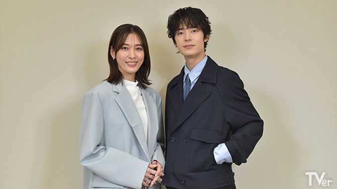 Two actors stand side by side in a studio setting with a light beige background. The woman on the left wears a light blue blazer over a white top and has shoulder-length dark hair. The man on the right wears a dark blue suit jacket, white shirt, blue tie, and has curly dark hair. A TVer logo appears in the bottom right corner.