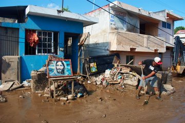 First image shows a young woman with long dark hair wearing a gray t-shirt and denim shorts sitting on a muddy street surrounded by floodwater piles of trash plastic containers and debris near metal gates and buildings in an urban alley. Second image is an aerial view of a flooded neighborhood with brown water covering the ground cars partially submerged laundry hanging on lines trees and white multi-story buildings along the street with scattered debris. Third image depicts a yellow two-story house partially submerged in floodwater with green trees around scattered trash boxes and plastic bottles floating in the muddy water. Fourth image features a man in a black cap and t-shirt using a shovel to clear mud and debris near a blue house with a painting of Jesus on a wooden stand and scattered wood scraps in the foreground.