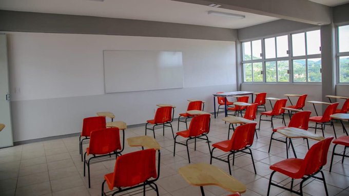 Empty classroom interior with multiple red plastic chairs and attached wooden desks arranged in rows facing a large blank whiteboard on a white wall. Large windows on the right side allow natural light and reveal a view of green hills outside. Gray walls and tiled floor complete the simple educational setting.