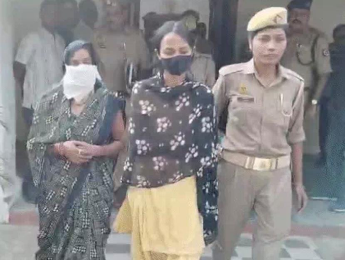Three women in traditional Indian attire including saris and dupattas one wearing a black printed shawl over yellow skirt another in green sari with white mask third in maroon sari with white mask flanked by uniformed male police officers with caps and badges standing outside building entrance with crowd of men in background on steps.