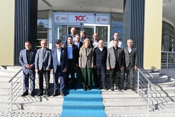 First image shows a group of men in suits seated around a large oval conference table in a modern room with beige walls, a projector screen, Turkish flag, and organizational banners; water bottles and flower arrangements are on the table; a portrait hangs on the wall. Second image depicts a group of about twelve people including men and women in formal attire standing on steps with a blue carpet at an entrance marked with a red 100 sign and crescent moon emblem, surrounded by glass doors and columns. Third image features men in suits around a rectangular table in a room with wooden paneling, Turkish flag, organizational emblems, flower centerpieces, and water bottles; a portrait is visible on the wall.