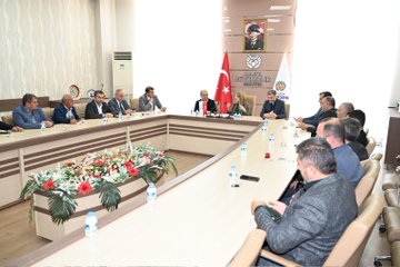 First image shows a group of men in suits seated around a large oval conference table in a modern room with beige walls, a projector screen, Turkish flag, and organizational banners; water bottles and flower arrangements are on the table; a portrait hangs on the wall. Second image depicts a group of about twelve people including men and women in formal attire standing on steps with a blue carpet at an entrance marked with a red 100 sign and crescent moon emblem, surrounded by glass doors and columns. Third image features men in suits around a rectangular table in a room with wooden paneling, Turkish flag, organizational emblems, flower centerpieces, and water bottles; a portrait is visible on the wall.