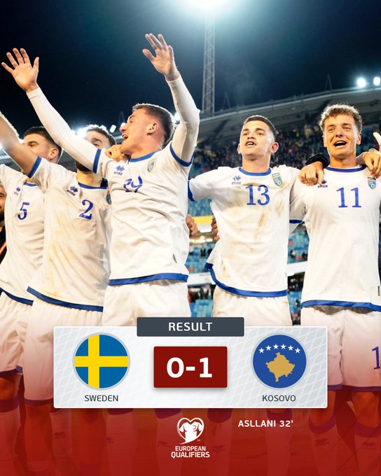 Group of male soccer players in white jerseys with blue accents and numbers 5 13 11 24 celebrating a victory by raising their arms high under bright stadium floodlights at night with a blurred crowd in the background and a graphic overlay at the bottom showing the Swedish flag a red 0-1 scoreline the Kosovo flag and text Result Sweden 0-1 Kosovo Asllani 32 WCQ