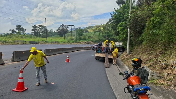 Several workers wearing yellow vests and helmets stand near orange traffic cones and barriers on a roadside with a dirt shoulder and asphalt pavement. A truck loaded with gravel is positioned nearby, and some workers handle tools or materials. An orange motorcycle is parked on the side, with a rider in helmet visible. Green hills and utility poles line the background under a partly cloudy sky.