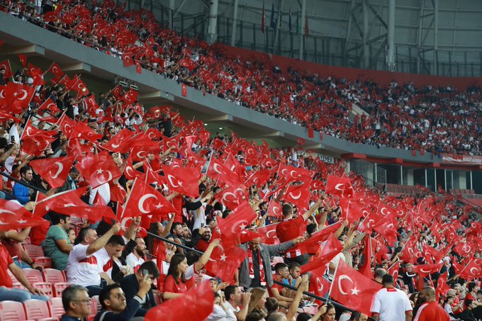 A large crowd of spectators fills the stands of a stadium, many wearing red clothing and waving numerous red flags with white crescents and stars, creating a sea of red in the seating areas under bright stadium lights.