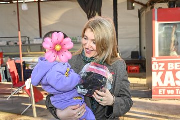 First image shows a blonde woman in a gray suit and green scarf holding a toddler boy in a purple hooded sweatshirt with a large pink flower toy attached to his hood, standing outdoors near red chairs and tables under a tent structure with shelves and cans in the background. Second image depicts the same woman leaning over a table in a modern cafe with wooden decor and plants on walls, assisting a young girl with curly hair in a white sweater eating from a white bowl of food, with another woman in striped shirt sitting nearby and two men in dark suits standing behind. Third image captures the woman in gray suit standing beside the young girl at the cafe table with plates and drinks, surrounded by similar decor including hanging wreaths and abstract wall art, with an older woman in blue and a man in suit nearby. Fourth image features the blonde woman in gray suit holding the toddler boy with the pink flower toy, standing near market shelves with red chairs and a sign reading OZ BEKAS EST in Turkish, with bags and containers visible.