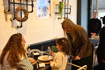 First image shows a blonde woman in a gray suit and green scarf holding a toddler boy in a purple hooded sweatshirt with a large pink flower toy attached to his hood, standing outdoors near red chairs and tables under a tent structure with shelves and cans in the background. Second image depicts the same woman leaning over a table in a modern cafe with wooden decor and plants on walls, assisting a young girl with curly hair in a white sweater eating from a white bowl of food, with another woman in striped shirt sitting nearby and two men in dark suits standing behind. Third image captures the woman in gray suit standing beside the young girl at the cafe table with plates and drinks, surrounded by similar decor including hanging wreaths and abstract wall art, with an older woman in blue and a man in suit nearby. Fourth image features the blonde woman in gray suit holding the toddler boy with the pink flower toy, standing near market shelves with red chairs and a sign reading OZ BEKAS EST in Turkish, with bags and containers visible.