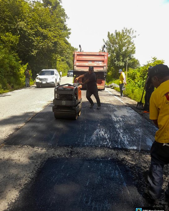 First image shows several workers in yellow shirts and helmets around a red dump truck unloading asphalt onto a road section surrounded by green vegetation with one worker holding a tool near the fresh black pavement patch. Second image depicts workers operating a small yellow roller machine compacting wet asphalt on the road beside the dump truck and a white vehicle parked nearby in a lush roadside setting. Third image captures a group of workers in safety gear standing near a damaged road area with potholes and debris on a hillside road lined with trees under a cloudy sky. Fourth image illustrates a long stretch of rural road with a fresh asphalt patch being applied visible in the foreground headlights from an approaching vehicle illuminating the scene amid dense green foliage.