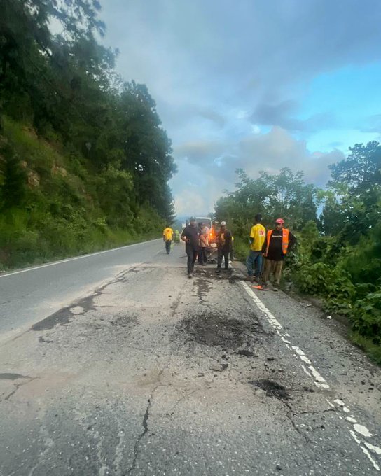 First image shows several workers in yellow shirts and helmets around a red dump truck unloading asphalt onto a road section surrounded by green vegetation with one worker holding a tool near the fresh black pavement patch. Second image depicts workers operating a small yellow roller machine compacting wet asphalt on the road beside the dump truck and a white vehicle parked nearby in a lush roadside setting. Third image captures a group of workers in safety gear standing near a damaged road area with potholes and debris on a hillside road lined with trees under a cloudy sky. Fourth image illustrates a long stretch of rural road with a fresh asphalt patch being applied visible in the foreground headlights from an approaching vehicle illuminating the scene amid dense green foliage.