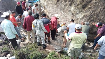First image shows multiple people including uniformed personnel in camouflage and civilians in casual clothing gathered around a large rocky slope with a massive boulder partially dislodged holding shovels and tools preparing to clear the obstruction. Second image depicts a group of individuals surrounding an enormous gray rock embedded in the ground amidst green vegetation with some using shovels to dig around its base while others stand nearby observing. Third image captures uniformed members in tactical gear and locals near collapsed structures and debris piles in a hilly area with scattered rocks and a red container visible on the ground. Fourth image illustrates personnel in camouflage uniforms assisting with blue tarps and wooden debris in a forested clearing with tents and damaged materials suggesting ongoing recovery efforts.