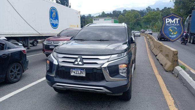 Gray Mitsubishi SUV with license plate P431NT parked on a road with traffic barriers, surrounded by other vehicles including a white truck displaying Chiquita logo, a red car, and a black sedan, in an urban setting with greenery and a police checkpoint sign visible nearby.
