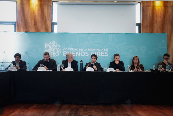 Group of suited professionals including men and women seated at a long conference table in a modern room with wooden floors and walls, a projector screen in the background, and a large blue banner displaying Gobierno de la Provincia de Buenos Aires logo, with water glasses, notebooks, and microphones on the table, one person speaking into a microphone.