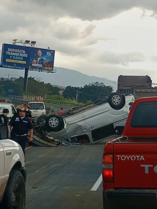 Overturned white vehicle lies on its side across a highway lane amid traffic, with police officers in blue uniforms standing nearby examining the wreckage. A red Toyota pickup truck is parked close by, and emergency response vehicles are visible. A blue billboard advertising Banco Industrial deposits and transfers stands prominently in the background, with mountains and cloudy skies visible beyond the road and surrounding greenery.