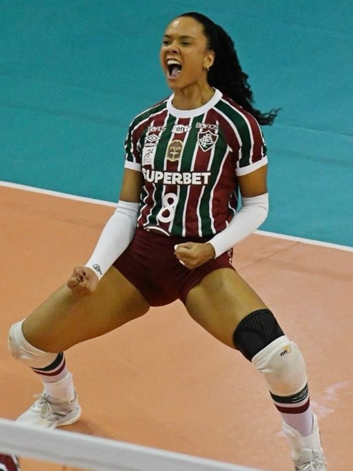 A female volleyball player with long braided hair wearing a green and white Superbet uniform jersey numbered 8, maroon shorts, white knee pads, and red and white socks stands triumphantly on an indoor blue court with arms raised in victory pose, mouth open in excitement, during a game setting.