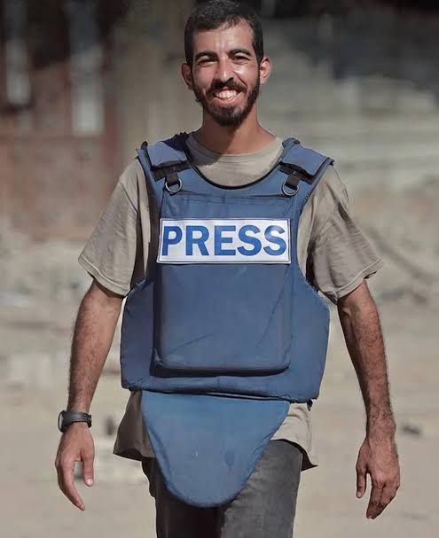 A man with short dark hair and a beard smiles while wearing a blue bulletproof vest labeled PRESS in white letters over a light gray t-shirt and dark pants in an outdoor urban setting with buildings and debris in the background.