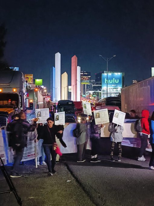 Nighttime urban scene on a road with multiple semi-trucks and cars stopped in traffic congestion, illuminated by streetlights and colorful high-rise tower structures in the background including a prominent red and white building with Hulu signage, groups of people standing on the asphalt holding white protest signs raised above their heads, some wearing hoodies and casual clothing, one person in a red jacket visible, overall atmosphere of a blockade protest with vehicles halted.