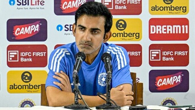 Gautam Gambhir sits at a press conference table wearing a blue shirt with white stripes, speaking into two microphones, with a serious expression, surrounded by sponsor banners including IDFC First Bank, SBI Life, Campa, Atomberg, and Dream11 on a colorful backdrop.
