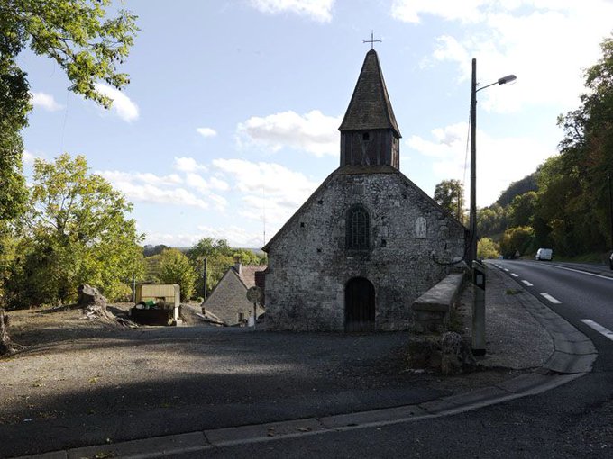 Stone chapel with pointed steeple and cross on top stands beside a road in a rural area surrounded by trees and hills under a partly cloudy sky a sign on the door indicates it is closed and there is a stone wall nearby.