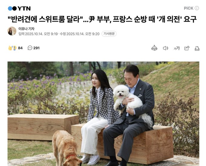 A man in a gray suit and a woman in a black-and-white checkered coat sit on wooden benches outdoors with green plants and flowers in the background. The woman holds a small white dog on her lap. A golden retriever sits nearby on the ground. The scene appears relaxed in a park-like setting.