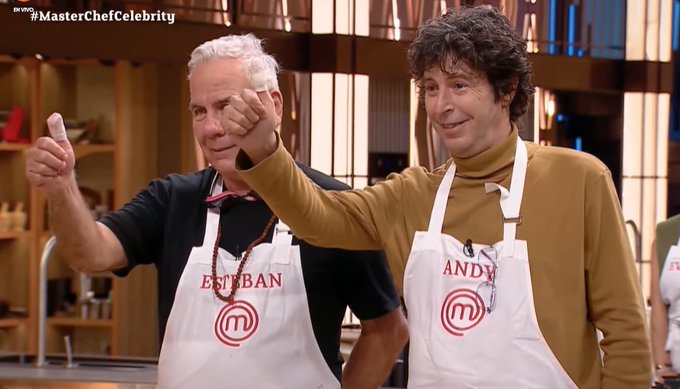 Two men stand in a modern kitchen setting on a TV show stage, wearing white aprons labeled with MasterChef Celebrity logo. The older man on the left has white hair, wears a black shirt, and gives a thumbs up with his right hand. The younger man on the right has curly dark hair, wears a brown turtleneck sweater, and also gives a thumbs up with his left hand, both smiling. Background includes wooden shelves, red stools, and kitchen utensils.
