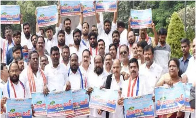Group of men and women in white shirts and saffron scarves stand outdoors holding blue banners with Telugu text including words like bandhu and reservations, some wearing traditional attire, trees in background.