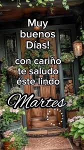 Rustic wooden door with glass panels in a garden surrounded by green vines, flower pots with pink flowers, a metal lantern hanging nearby, a stack of firewood beside it, and a stone path leading to the door. Overlaid text in green font reads Muyy buenos días con cariño te saludo este lindo and in purple cursive font Martes at the bottom.