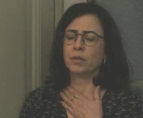 A woman with shoulder-length dark hair and round glasses has a contemplative expression, her mouth slightly open and eyes looking downward. She places her right hand gently on her chest. She wears a dark blouse with a subtle pattern and a ring on her finger. The background is a plain indoor wall with a door frame.