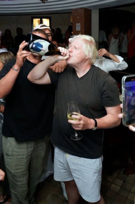 Boris Johnson with blonde hair in a black t-shirt holds a glass of wine in his right hand while a man in a black t-shirt and gray shorts embraces him from behind holding a large blue-labeled champagne bottle to Johnsons mouth as he drinks. A smartphone is visible in the foreground capturing the moment. A crowd of people in casual summer clothing stands around in a warmly lit indoor venue with arched ceiling and windows.