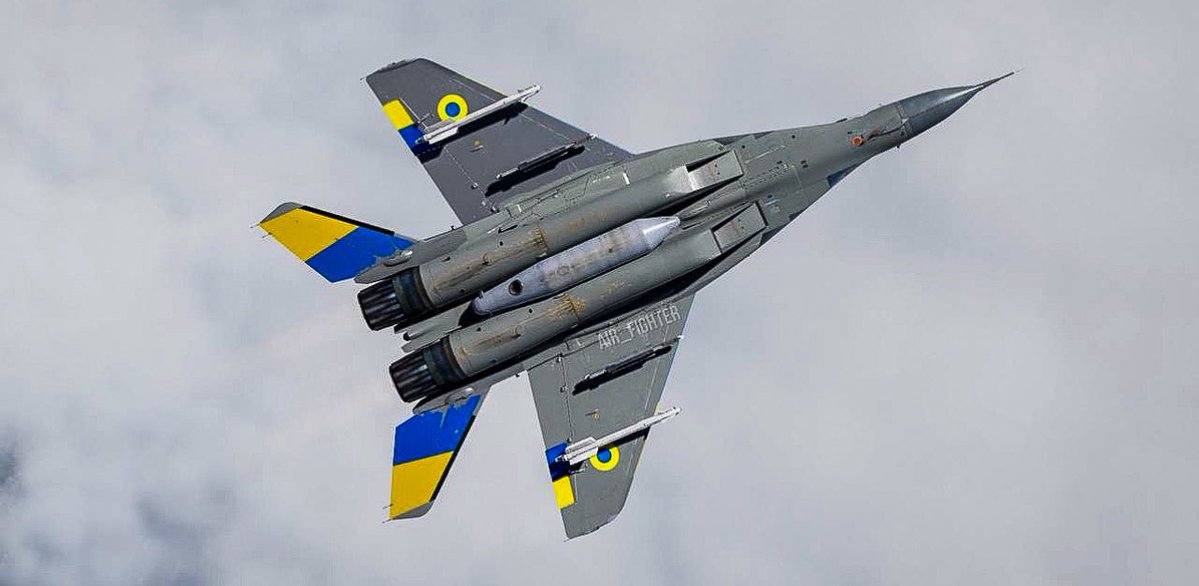 一架灰色的 MiG-29 战斗机在头顶飞过，其垂直安定面和机翼上有蓝色和黄色条纹，与乌克兰国旗颜色相匹配，从下方看，在部分多云的天空中，显示双引擎排气管和翼下挂载点。