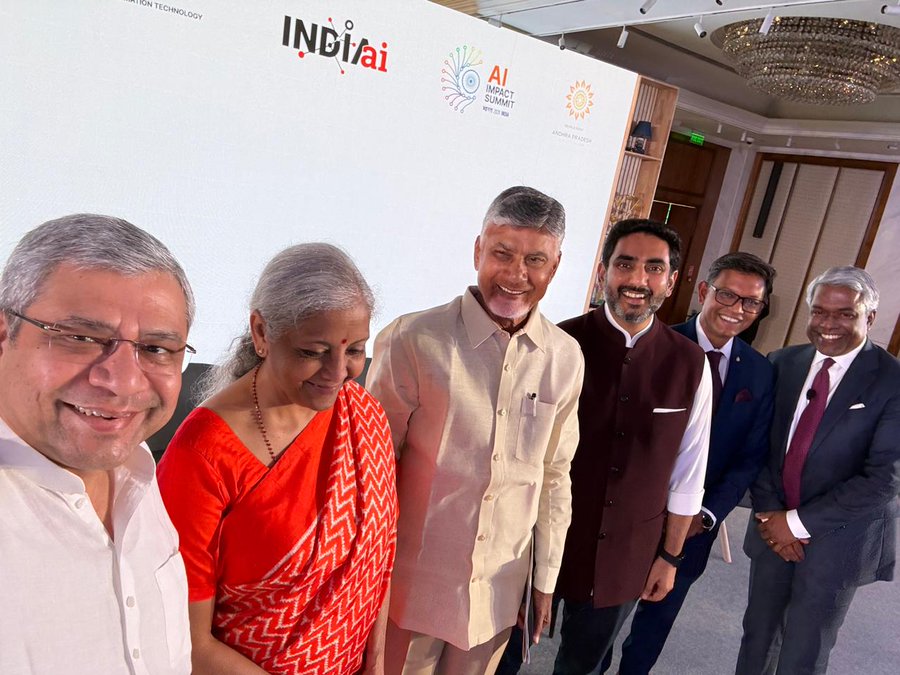 Group of six men and one woman in formal Indian attire including kurtas, sarees, suits, and vests stand smiling for a photo in an indoor venue with white walls, a chandelier, and backdrop banners displaying AI for India logo, G20 emblem, and event text. The individuals include an elderly man in cream kurta, a woman in red saree, a man in beige kurta, a younger man in maroon vest, another in blue suit, and two in suits with ties. Setting appears to be a conference or summit room with modern lighting and decor.