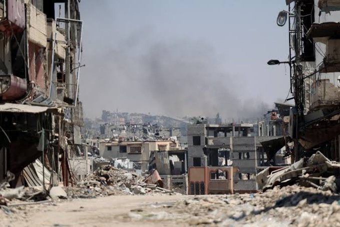 Devastated urban landscape in Shujaiya east of Gaza City showing multiple collapsed and damaged buildings with exposed interiors scattered rubble piles of debris narrow dirt paths between structures lingering smoke rising from various points hazy sky overhead streetlights and satellite dishes on remaining structures indicating recent bombing destruction