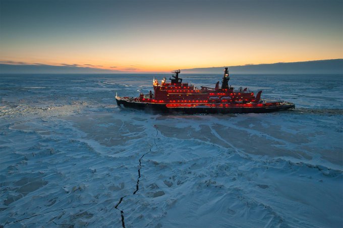 A red icebreaker ship with illuminated lights sails through vast expanses of blue-white Arctic ice under a twilight sky with orange sunset hues on the horizon. The vessel features multiple decks, antennas, and a prominent bow cutting through cracked ice floes. Surrounding scenery includes flat icy plains extending to the distant horizon with subtle cracks visible in the ice surface.