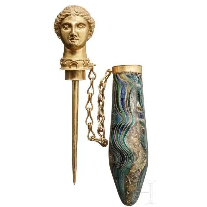 Frasco de perfume romano de vidrio con tapa dorada en forma de cabeza de mujer, aún unido por su cadena de oro. Con una larga punta en la parte inferior, el preciado perfume podía extraerse del recipiente de vidrio y aplicarse gota a gota. Fecha: siglo I d. C. aproximadamente.