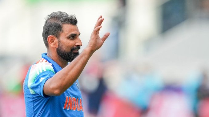 A man with short dark hair and beard wearing a blue India cricket jersey with orange accents stands on a field gesturing with his right hand raised palm forward while looking to the side blurred crowd and green background visible behind him.