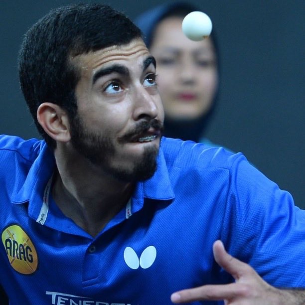 A man with short dark hair and a mustache wearing a blue collared shirt with Tenergy and Butterfly logos plays table tennis indoors, hitting a white ball mid-air with his paddle while extending his arm, a woman in a black hijab visible in the blurred background wearing blue athletic wear, another similar action shot from a different angle showing him swinging the paddle towards the ball with focused expression, and a third image of him in a blue Adidas shirt holding a red paddle in a ready stance on a table tennis court with Qatar Doubles Table Tennis Association banner behind.