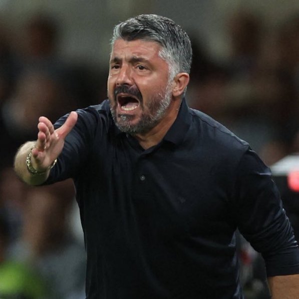 Man with gray hair and beard wearing black polo shirt gestures with open mouth and extended arm on soccer field surrounded by blurred stadium crowd.