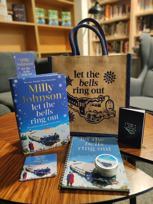 First image shows an open blue hardback book titled Let the Bells Ring Out by Milly Johnson with a signature inside held by a hand with a rainbow bracelet placed on a wooden table in a library setting with bookshelves and a lamp in the background Second image displays the same blue book standing on a wooden table next to a jute tote bag printed with Let the Bells Ring Out and train imagery a bookmark a notebook a coaster and a small tin container all arranged in a cozy room with bookshelves and an armchair