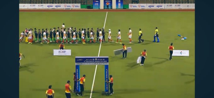 Group of male athletes in white jerseys with green accents and green jerseys with white accents stand in two lines on a green turf field shaking hands across a dividing line. Several officials in yellow shirts oversee the event. Banners reading ICSP and sponsor logos are visible on the field edges. An archway stands at the far end of the field with additional banners.