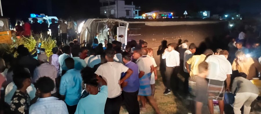 Nighttime scene on a road with an overturned white government bus lying on its side amid dirt and grass, surrounded by a large crowd of people in casual clothing including shirts and pants, some standing and others gathered near the vehicle, with nearby buildings, lights, and another bus visible in the background.