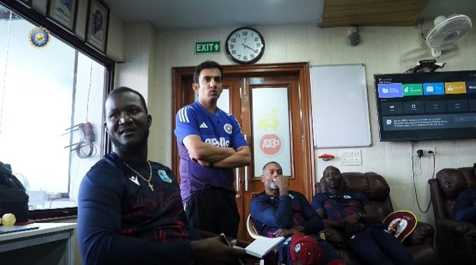 Several men of African descent and one with Indian features stand and sit in a well-lit room with wooden doors, a wall-mounted TV displaying a multicolored screen, a clock on the wall, a ceiling fan, large windows with blinds, framed pictures including an Indian flag emblem, and lounge chairs. One man wears a blue West Indies cricket jersey with arms crossed, another in a purple Chelsea football jersey holds a folder, others in dark clothing and a white shirt sit relaxed. The setting appears to be an office or lounge area.