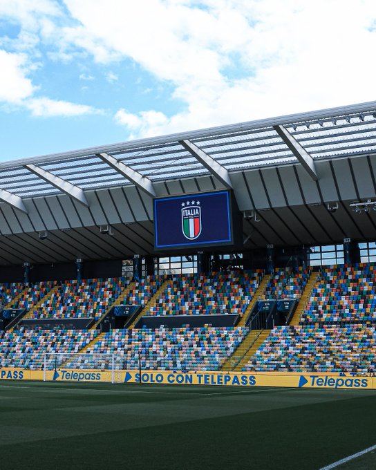 Stadium structure with metallic roof and large digital screen displaying the Italian national football team emblem in blue white and red colors against a clear sky with scattered clouds. Empty tiered seating in yellow and blue sections extends around the field. Green artificial turf pitch visible in foreground. Prominent advertising banners for Telepass along the lower edges in yellow with blue text reading TELEPASS SOLO CON TELEPASS.