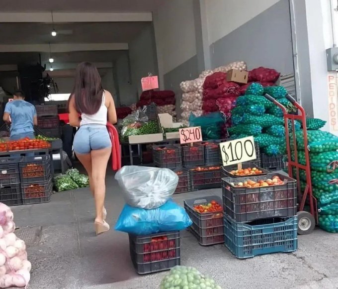 Stacks of green onions in red nets and crates priced at 100 reais sit on shelves next to tomatoes in orange containers priced at 10 reais. Garlic bulbs in pink bags and red peppers in crates are visible nearby. A woman with long dark hair wearing a white tank top and denim shorts bends over holding a red bag near the vegetable displays. Another woman in a blue shirt stands working at a table with produce. Blue plastic bags and carts with more onions are in the foreground under warehouse-style lighting.