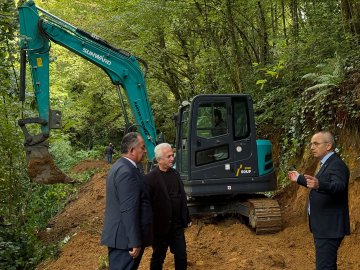 First image shows several men including one in suit gesturing near a white pickup truck in a forested area with green trees and dirt ground. Second image depicts three men in suits standing beside a green Sunward excavator on a dirt slope surrounded by trees and excavation dirt. Third image captures four men walking along a dirt path in a dense green forest with one in suit leading. Fourth image shows four men walking on a dirt trail amid green trees and foliage with one in suit holding glasses.